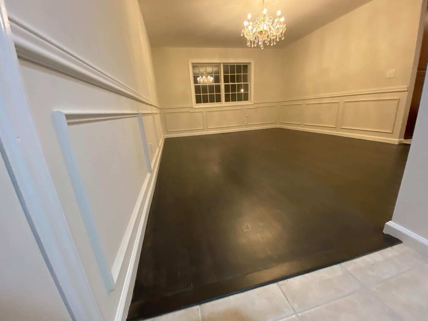 Empty dining room with dark hardwood floor, white wainscoting, chandelier, and window.