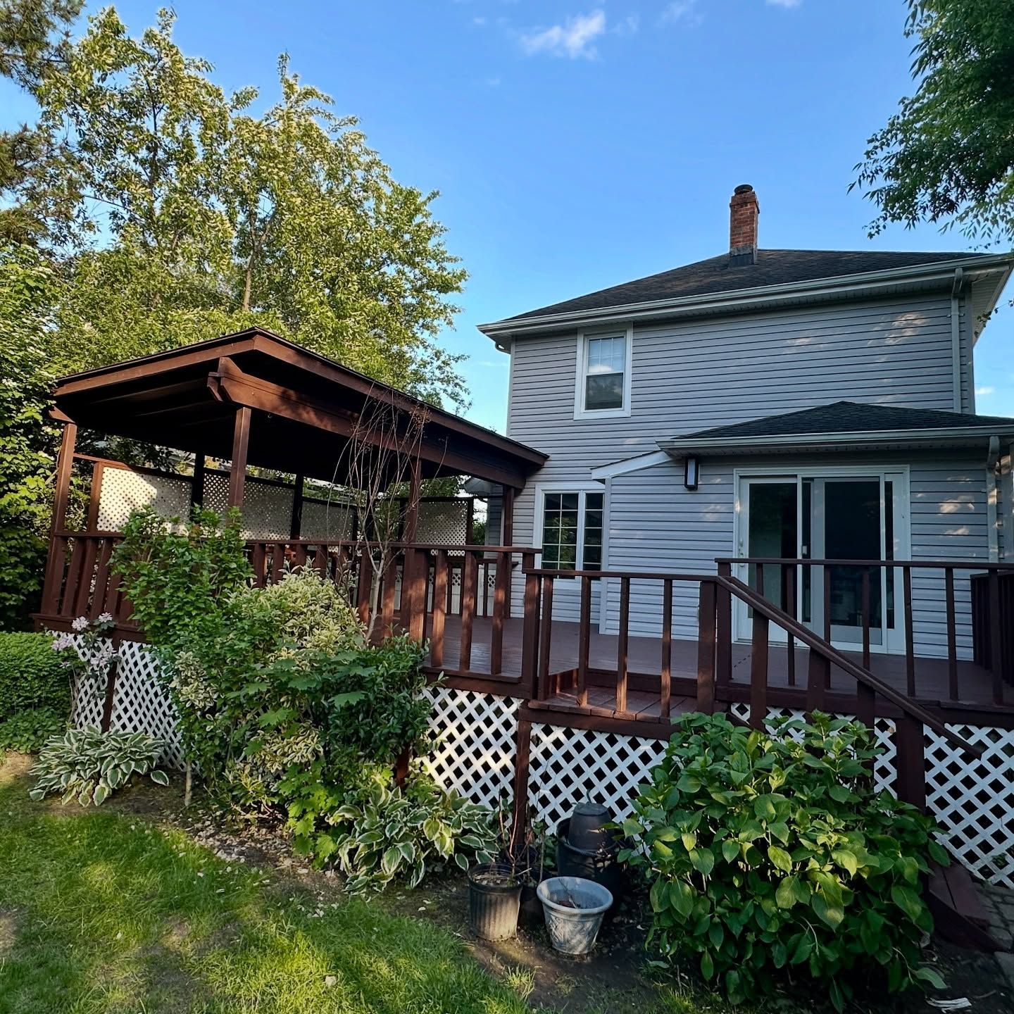 Backyard deck with gazebo, gray house, and lush green landscaping under trees