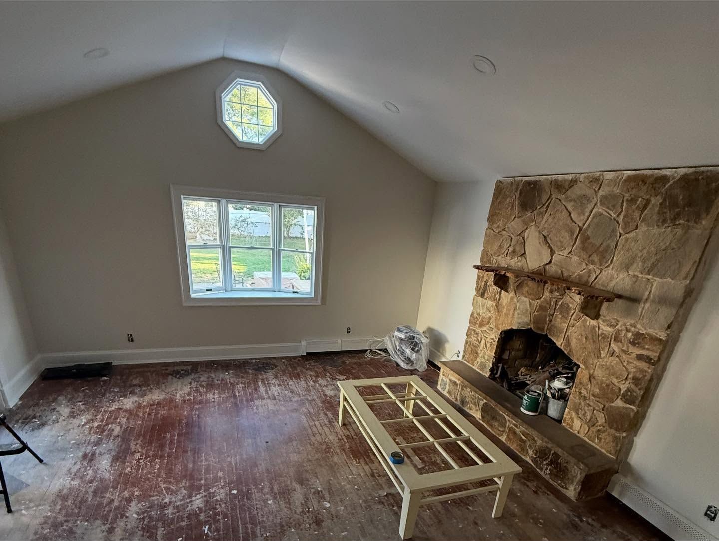 Empty room with beige walls, a window, octagonal clerestory window, and a stone fireplace under renovation