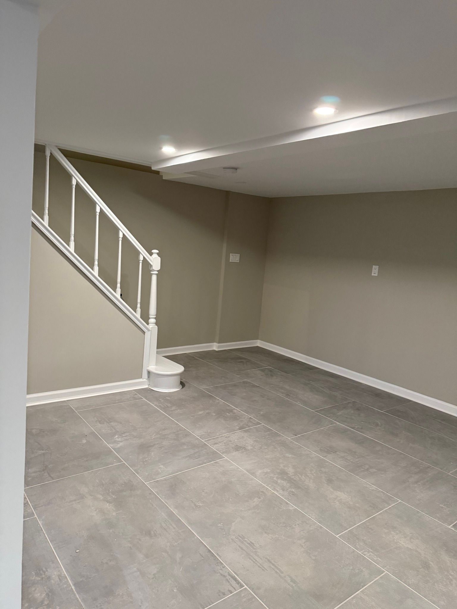 Empty beige basement room with tiled floor, white staircase, and recessed ceiling lights