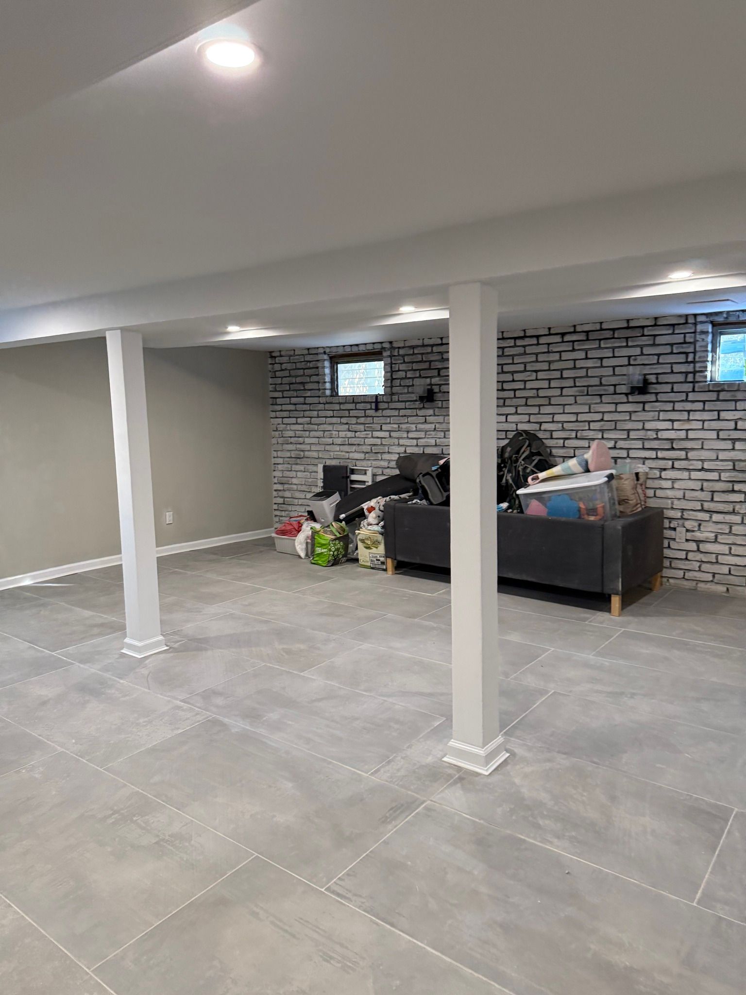 Spacious unfinished basement with gray tile floor, white support columns, and a brick accent wall with furniture.