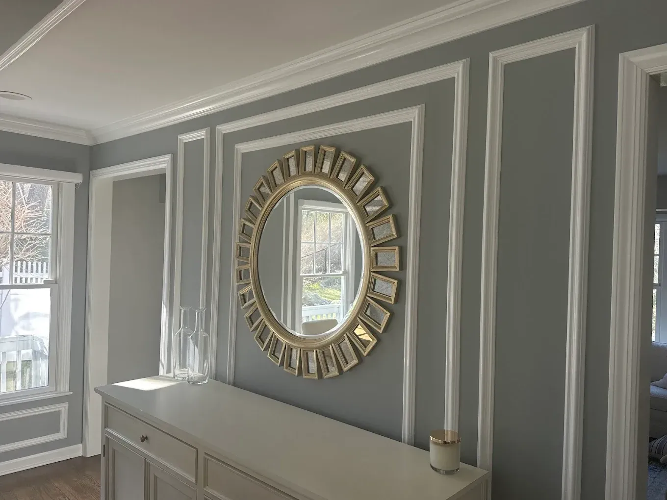 Gray paneled hallway with a round gold-framed mirror and white sideboard by a window