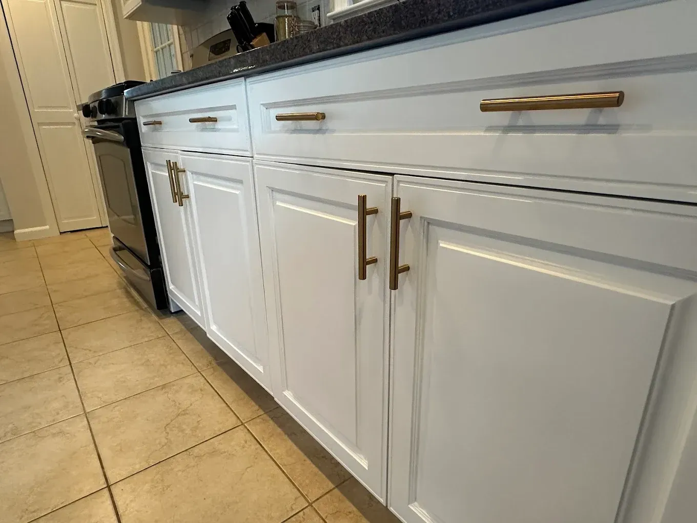 White kitchen cabinets with gold handles and black countertop beside a tiled floor.