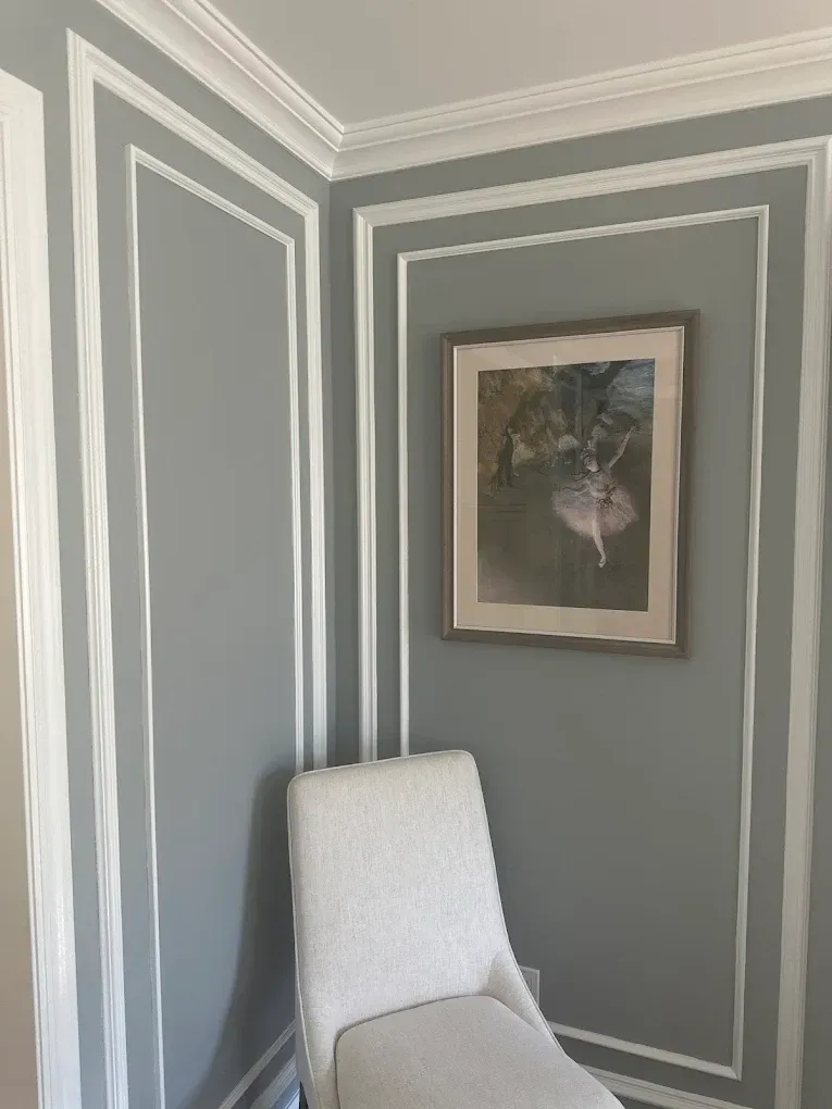 Gray corner wall with white trim, framed floral print, and a white upholstered chair below it.