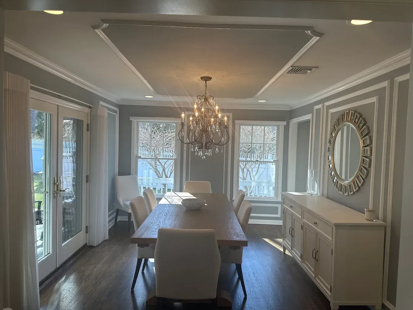 Elegant dining room with chandelier, long table, white chairs, and ornate wall mirror in gray tones