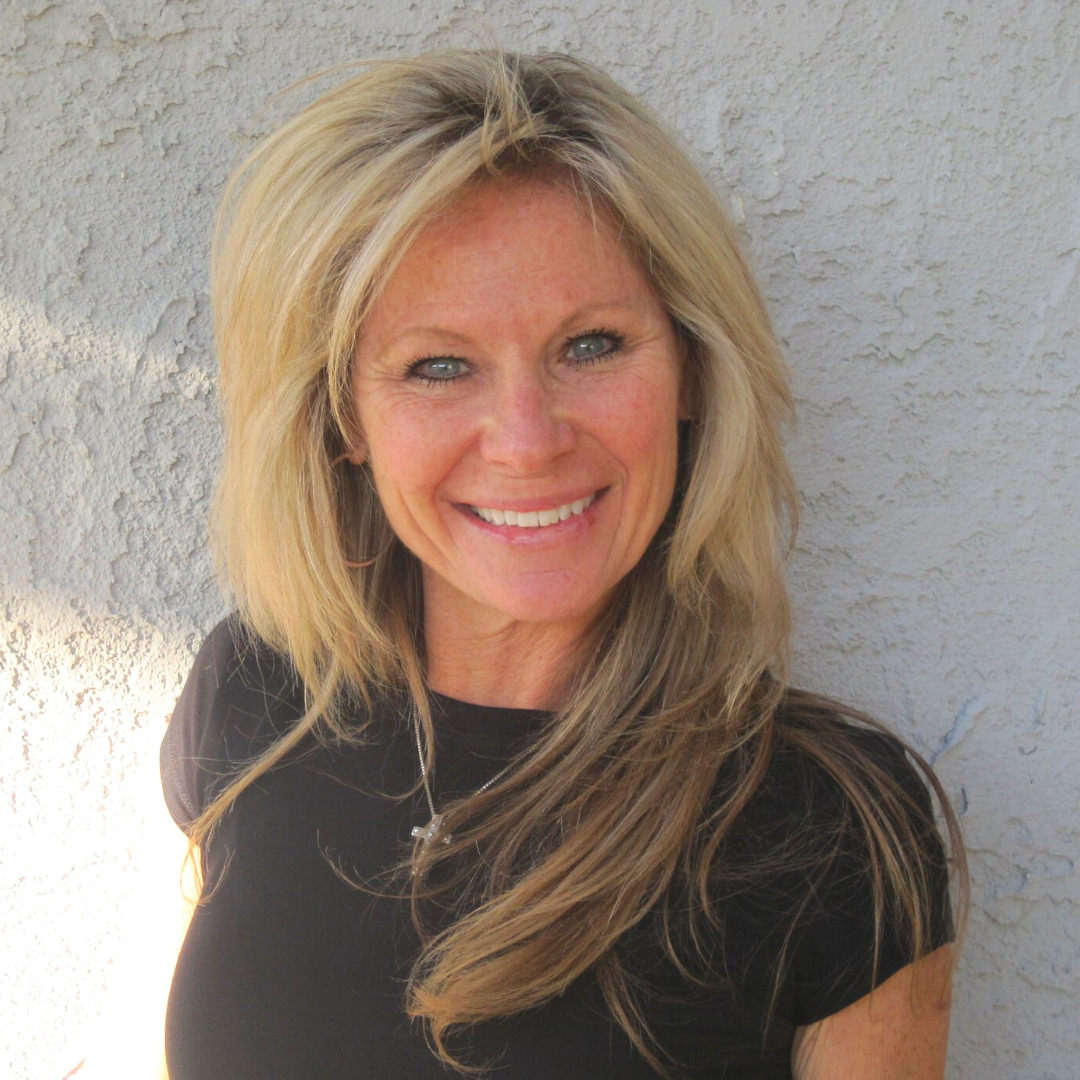 Blond woman with a big smile wearing a black shirt, standing against a light-colored wall.