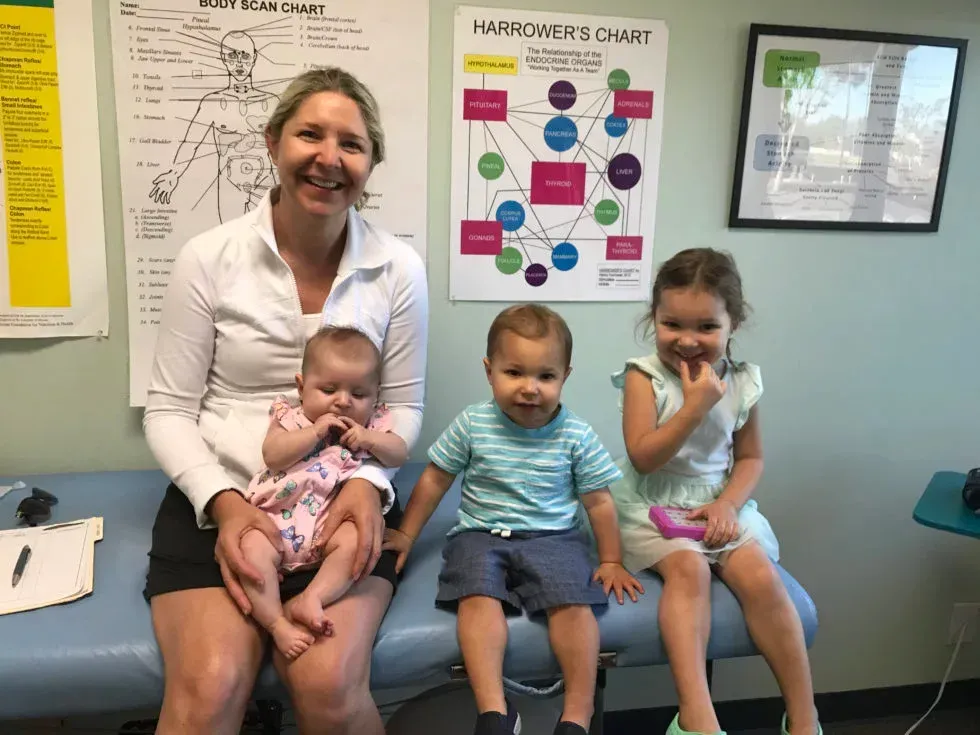 Woman with three children; sitting, smiling. Room with charts, desk. Children are looking and smiling.