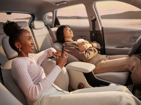Two women are sitting in the back seat of a car talking to each other. Kona