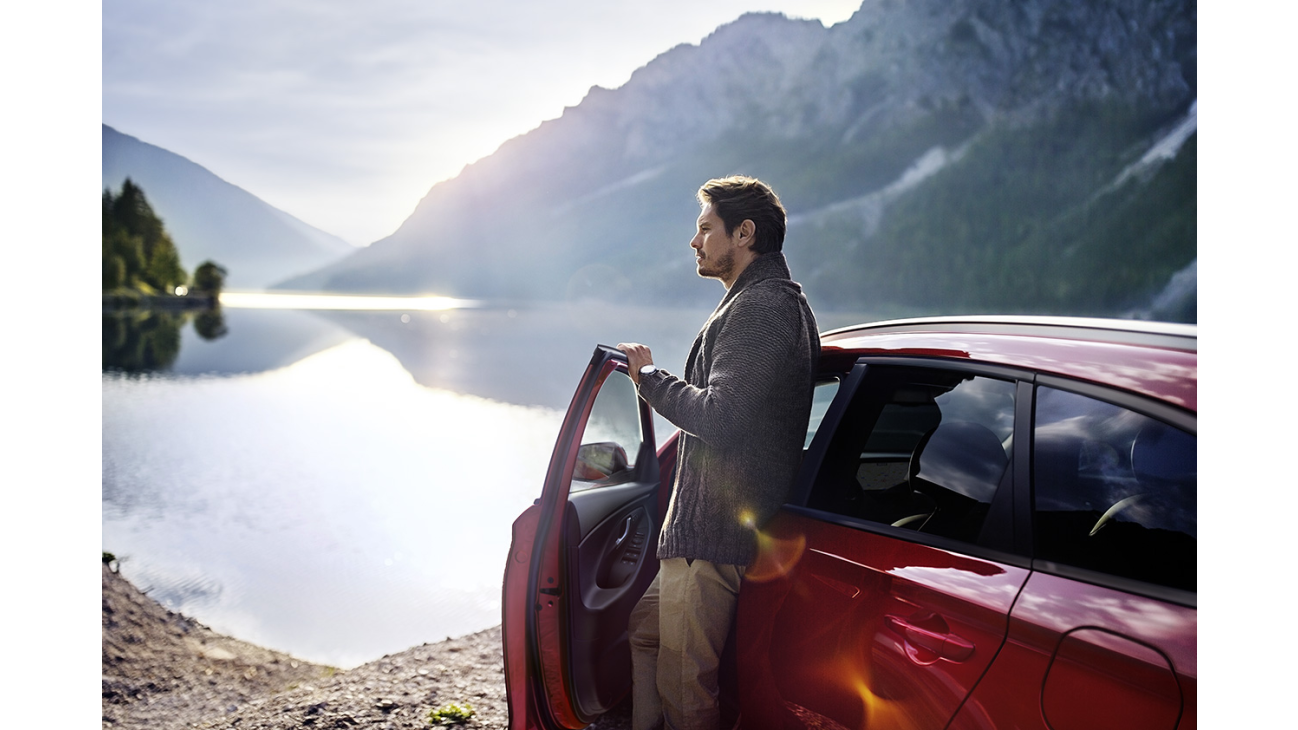 A man is standing next to a red car near a lake.