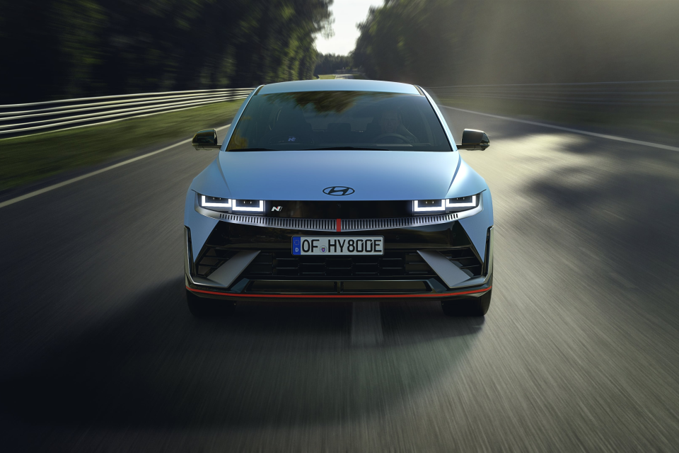 A blue car is driving down a road with trees in the background. Ioniq 5 N
