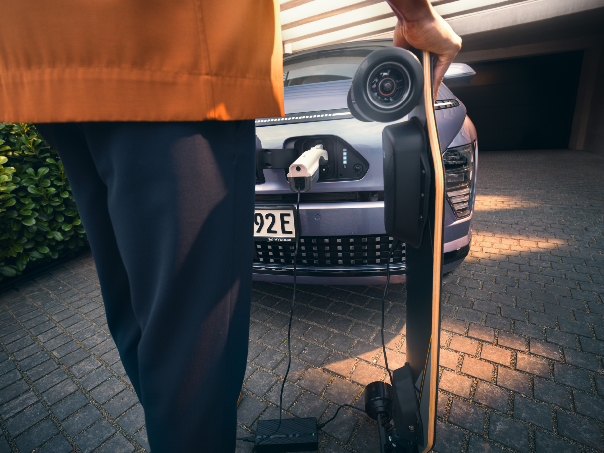 A person is holding a skateboard in front of a car that is being charged. Kona Electric