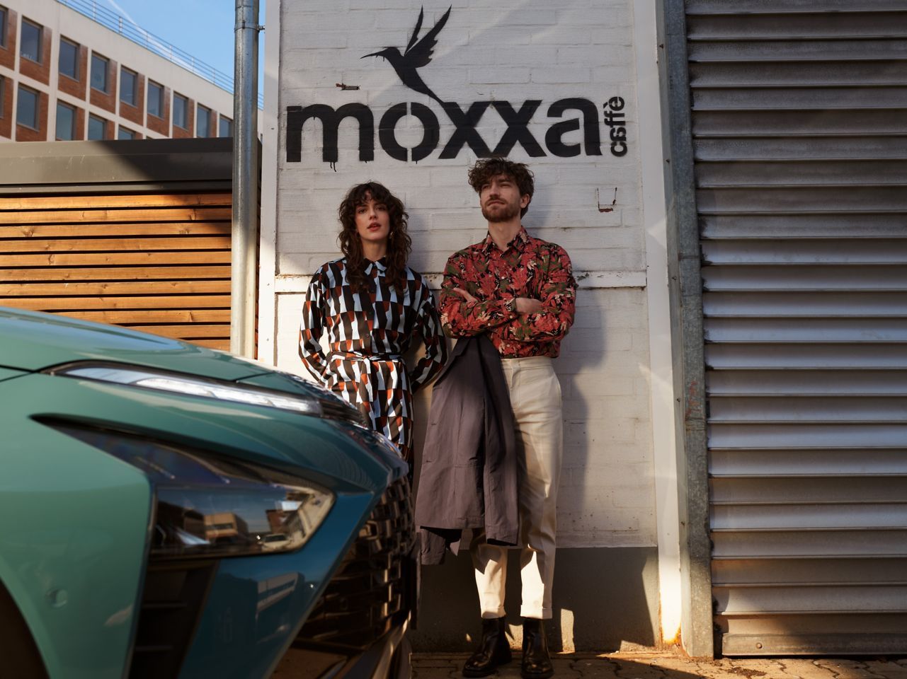 A man and a woman are standing next to a car in front of a moxxa sign. Bayon