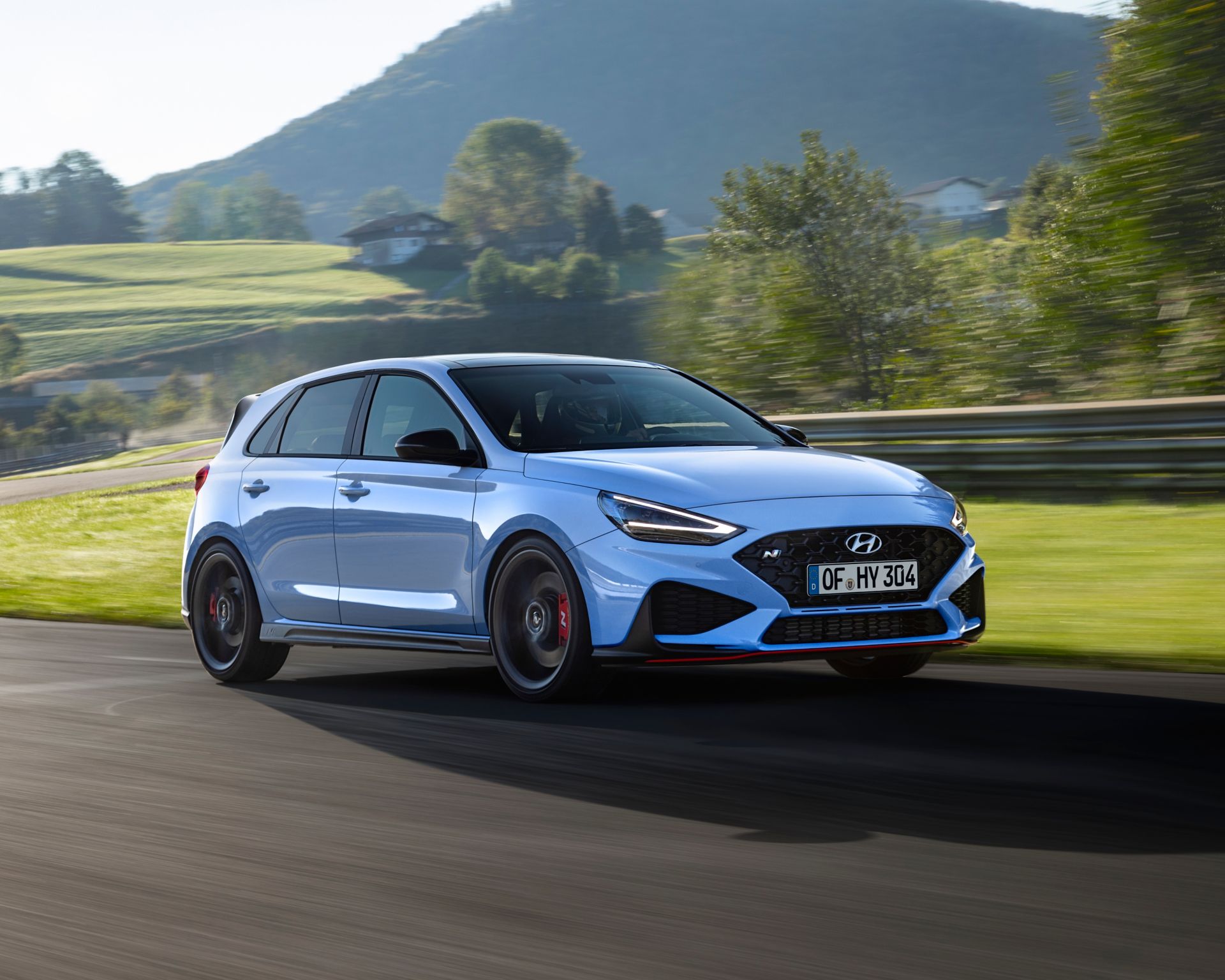 A blue car is driving down a road with mountains in the background. i30