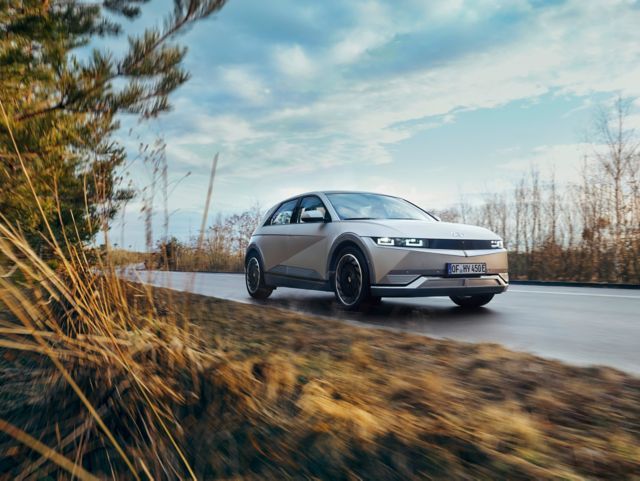 A white car is driving down a road next to tall grass. ioniq 5