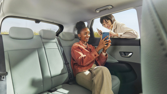 Two women are sitting in the back seat of a car looking at their phones. Kona