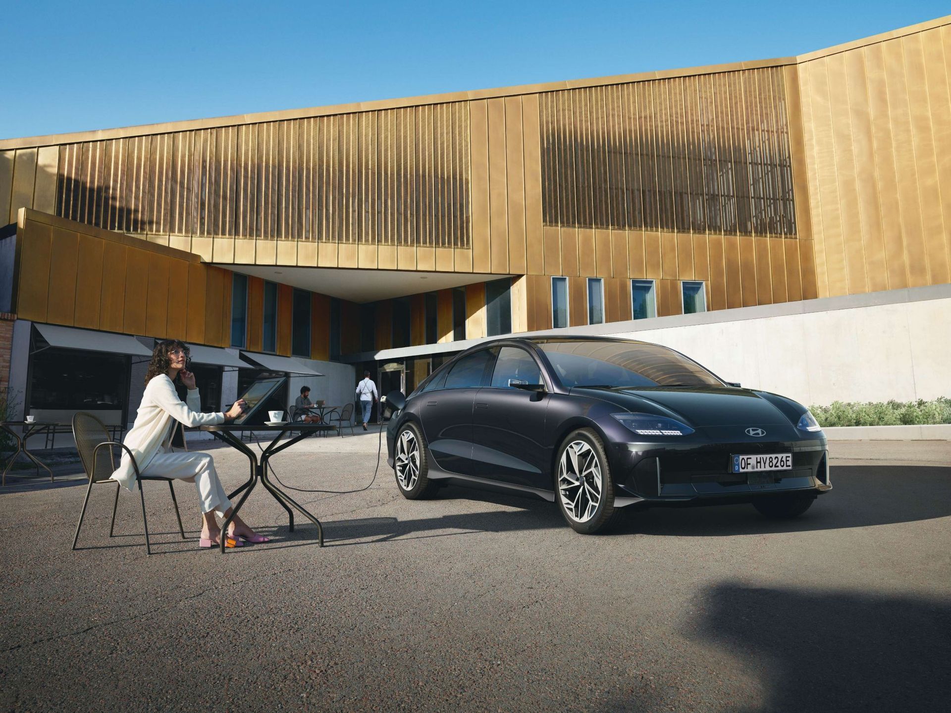 A woman is sitting at a table next to a car parked in front of a building. Ioniq 6