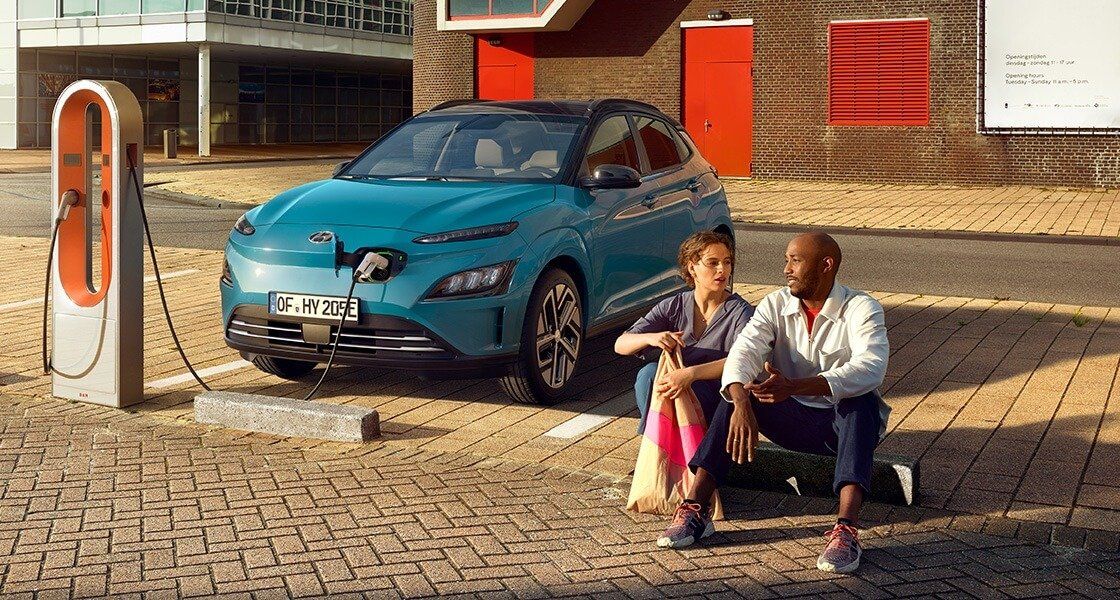 A man and a woman are sitting next to a blue electric car at a charging station.