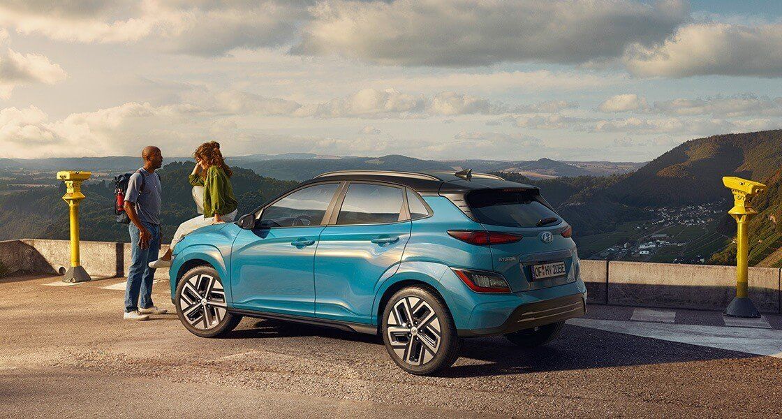 A man and a woman are standing next to a blue car on top of a hill.