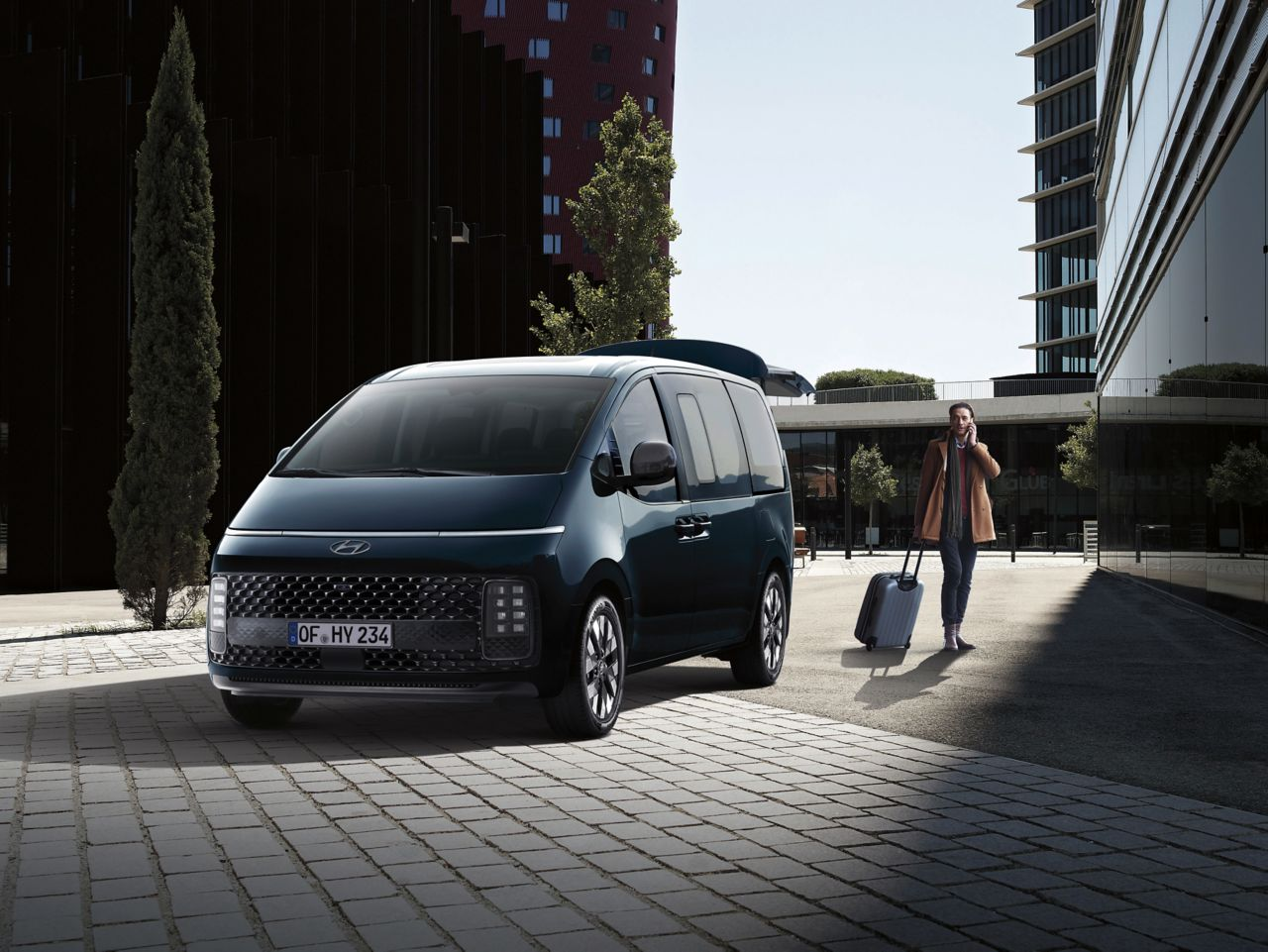 A man is walking towards a blue van with a suitcase. Staria