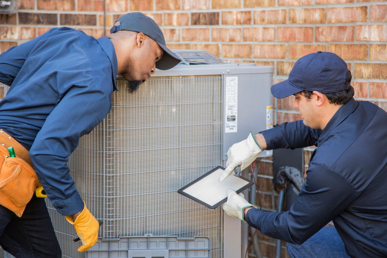 Two men are working on an air conditioner outside of a brick building.