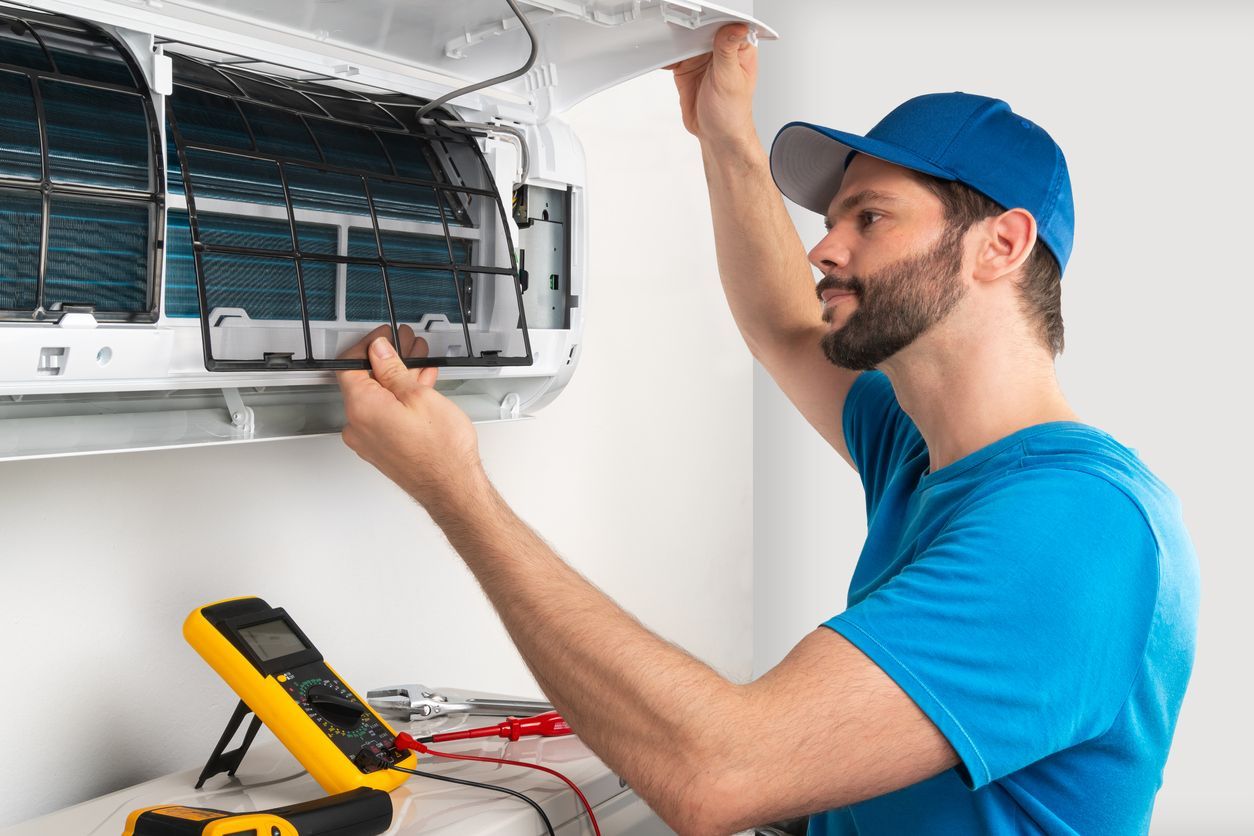 Technician in blue cap and shirt inspecting a wall-mounted air conditioner with tools nearby.