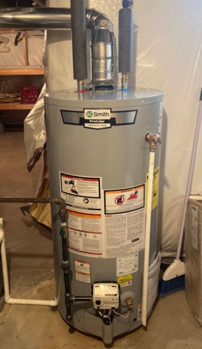 A water heater is sitting in a basement next to a broom.