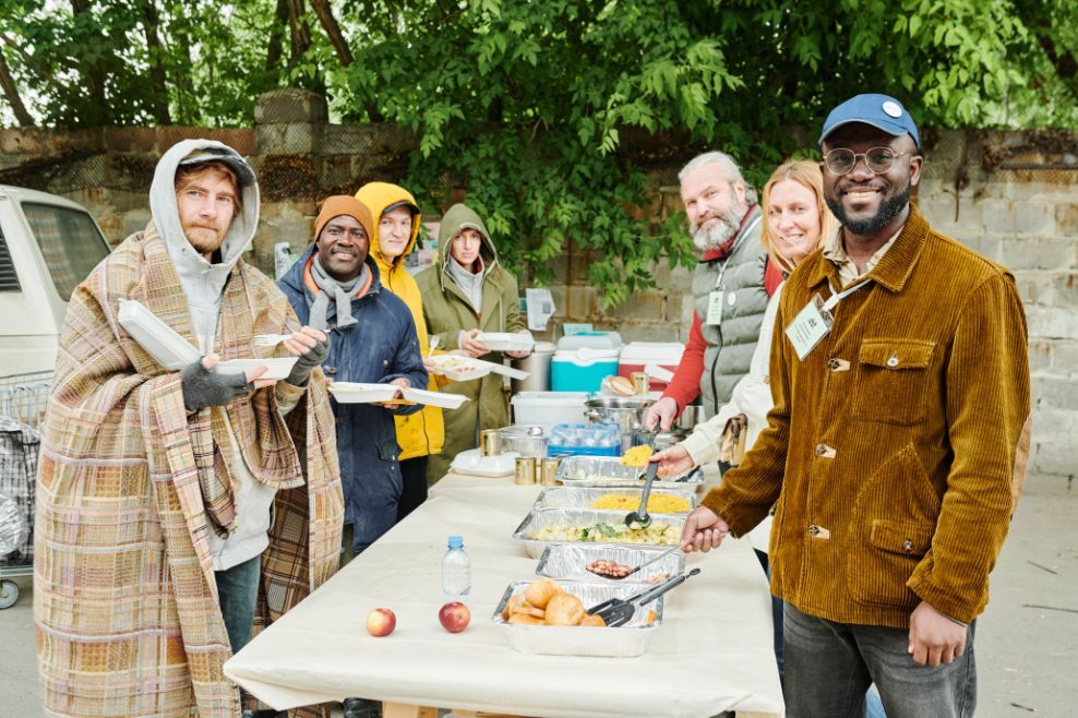 People serving food at an outdoor meal. A man smiles at the camera. Others reach for the food.