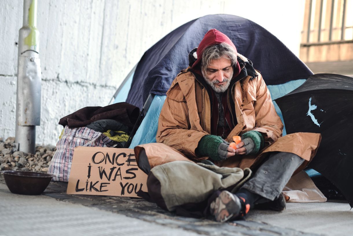 An unhoused man has a sign that reads,
