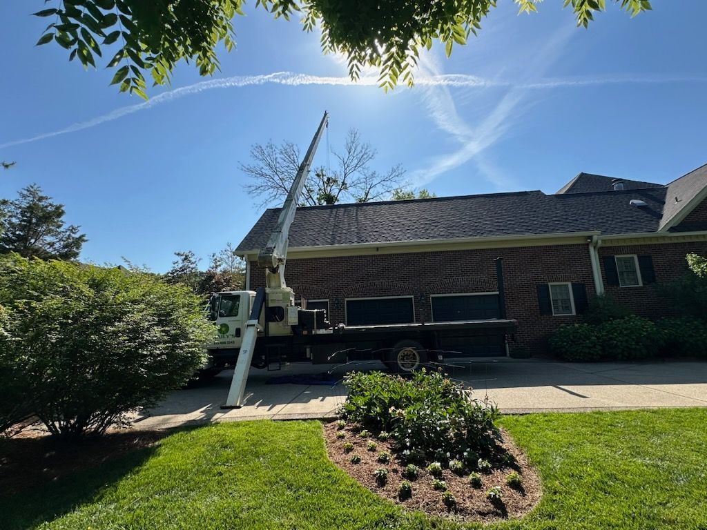 Bucket truck raised beside a brick house under a bright blue sky, with trees and a green lawn.