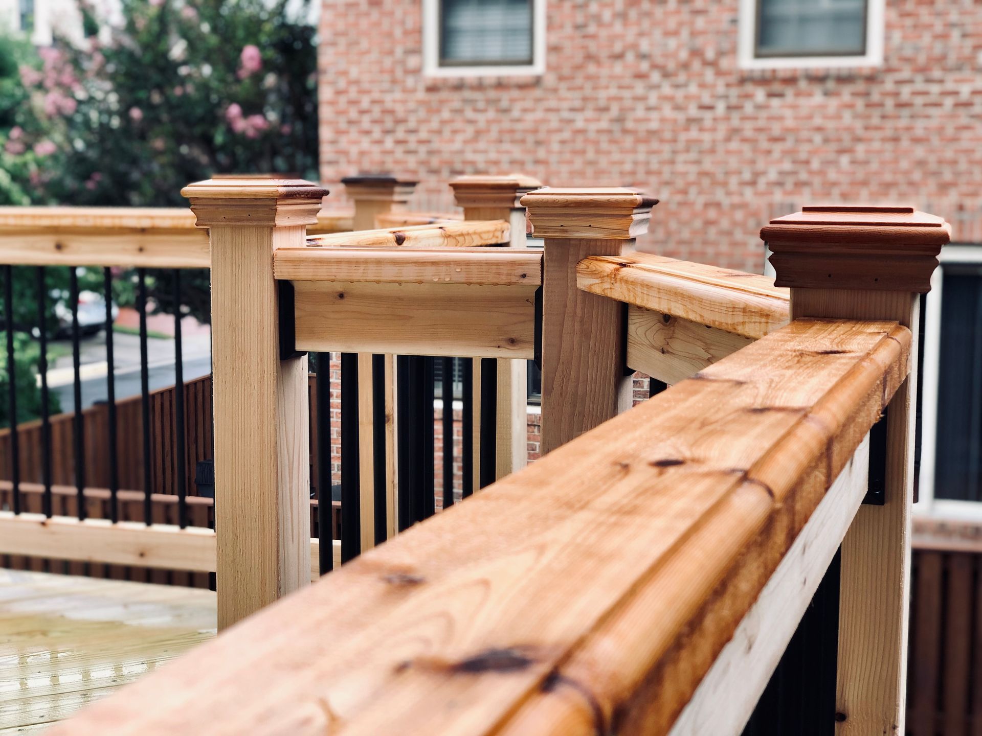 Wooden deck railing with capped posts; brown stained wood. Brick building background.