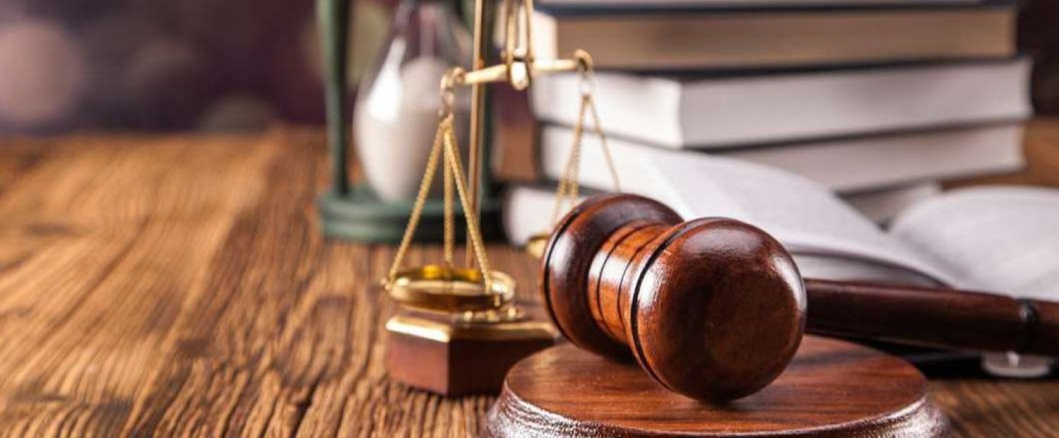 Wooden gavel and sound block next to a scale of justice, books, and an hourglass, on a wooden desk.