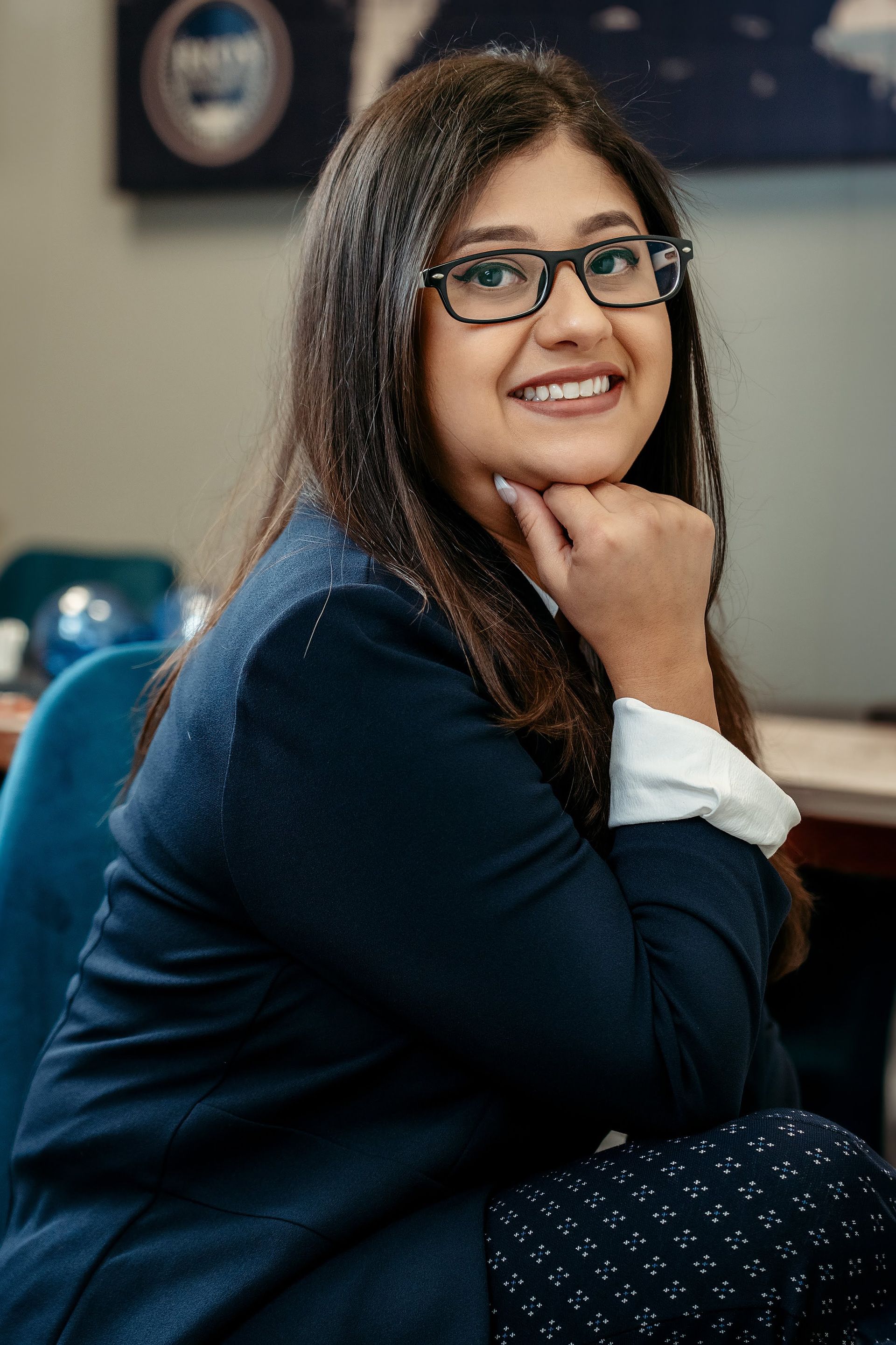 Woman with glasses smiles, resting chin on hand. She wears a blazer and sits in an office setting.