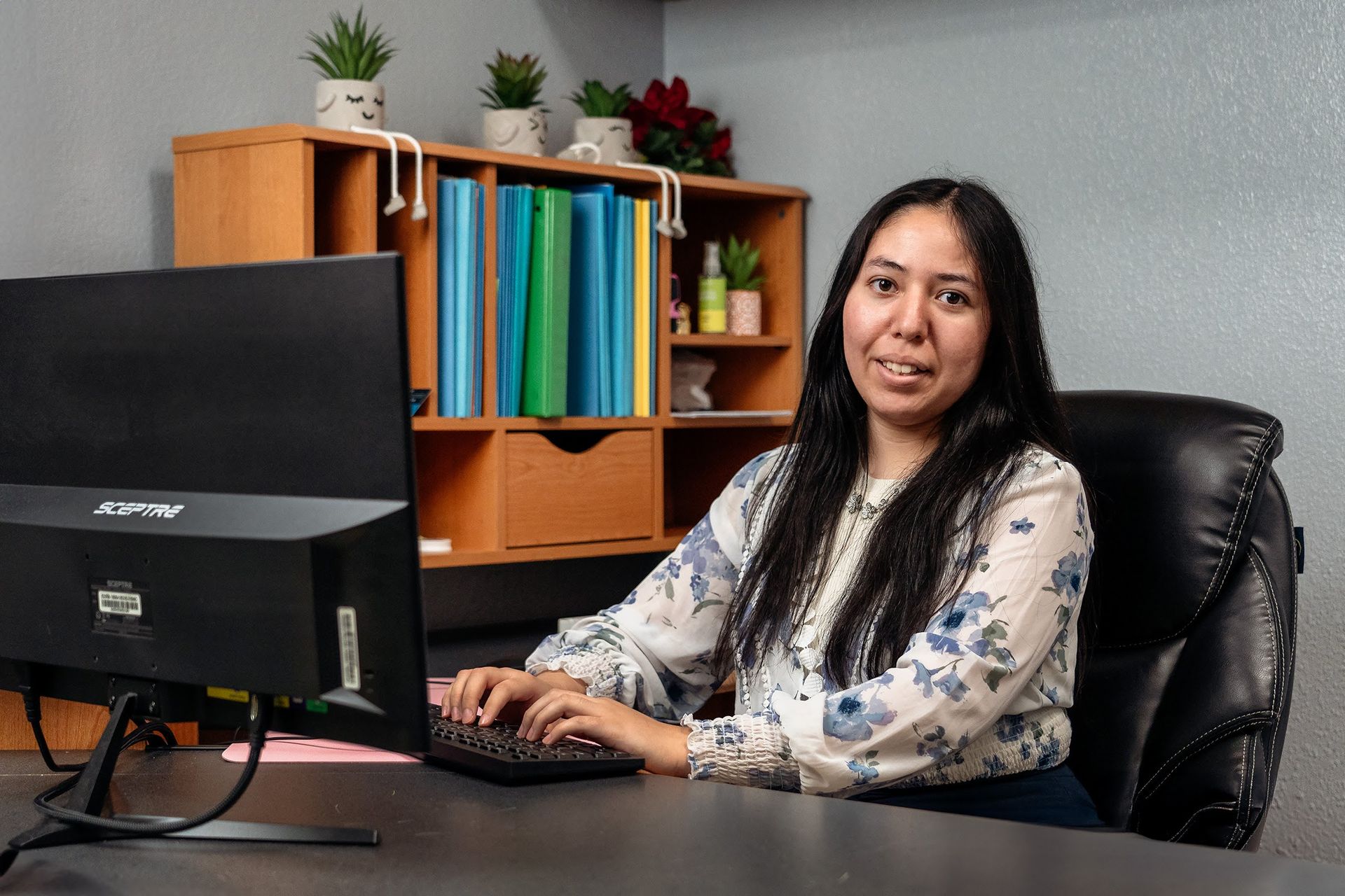 Woman at desk typing on keyboard, smiling; books, plants, and monitor in background.