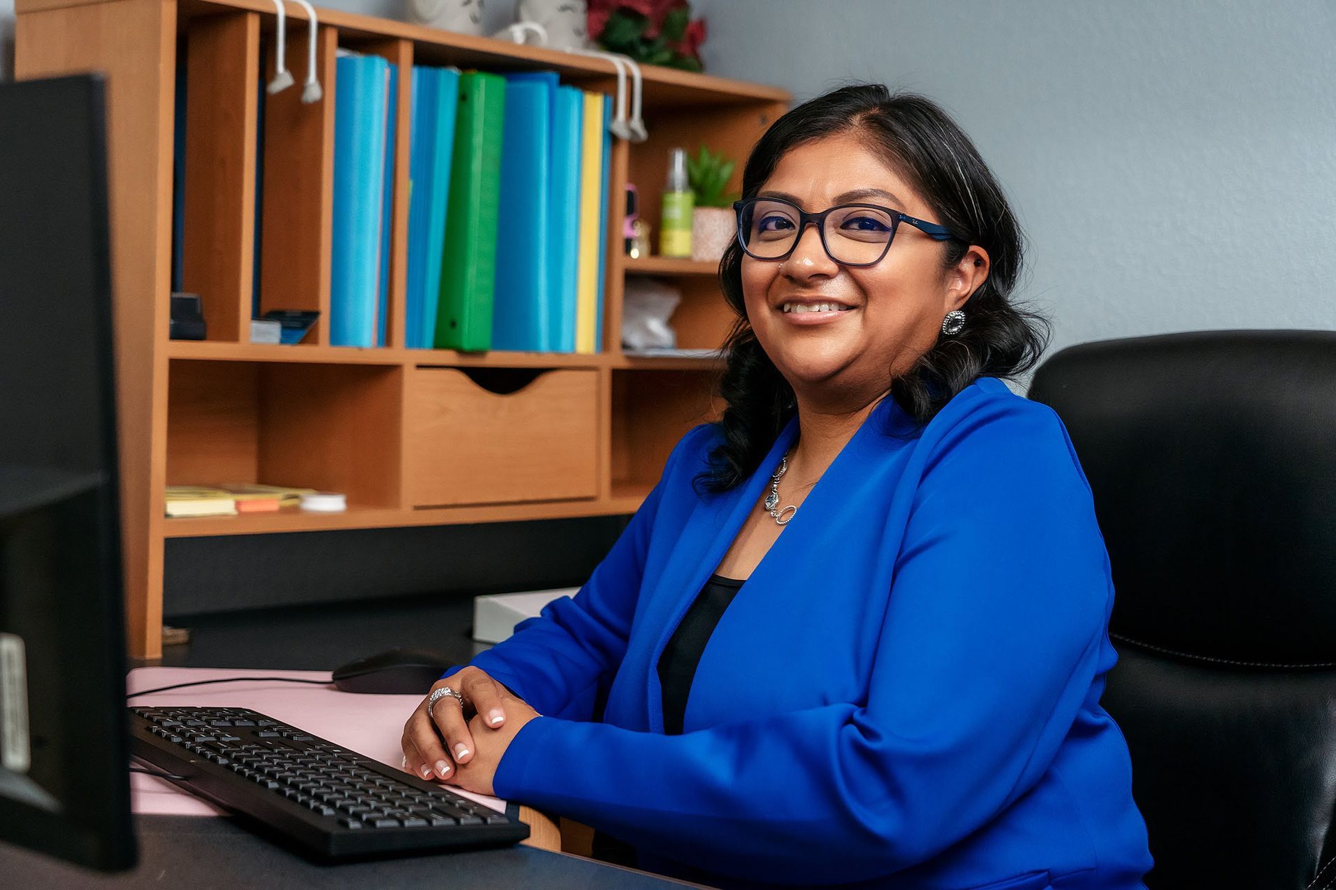 Woman with glasses smiles while sitting at a desk, wearing a blue blazer.
