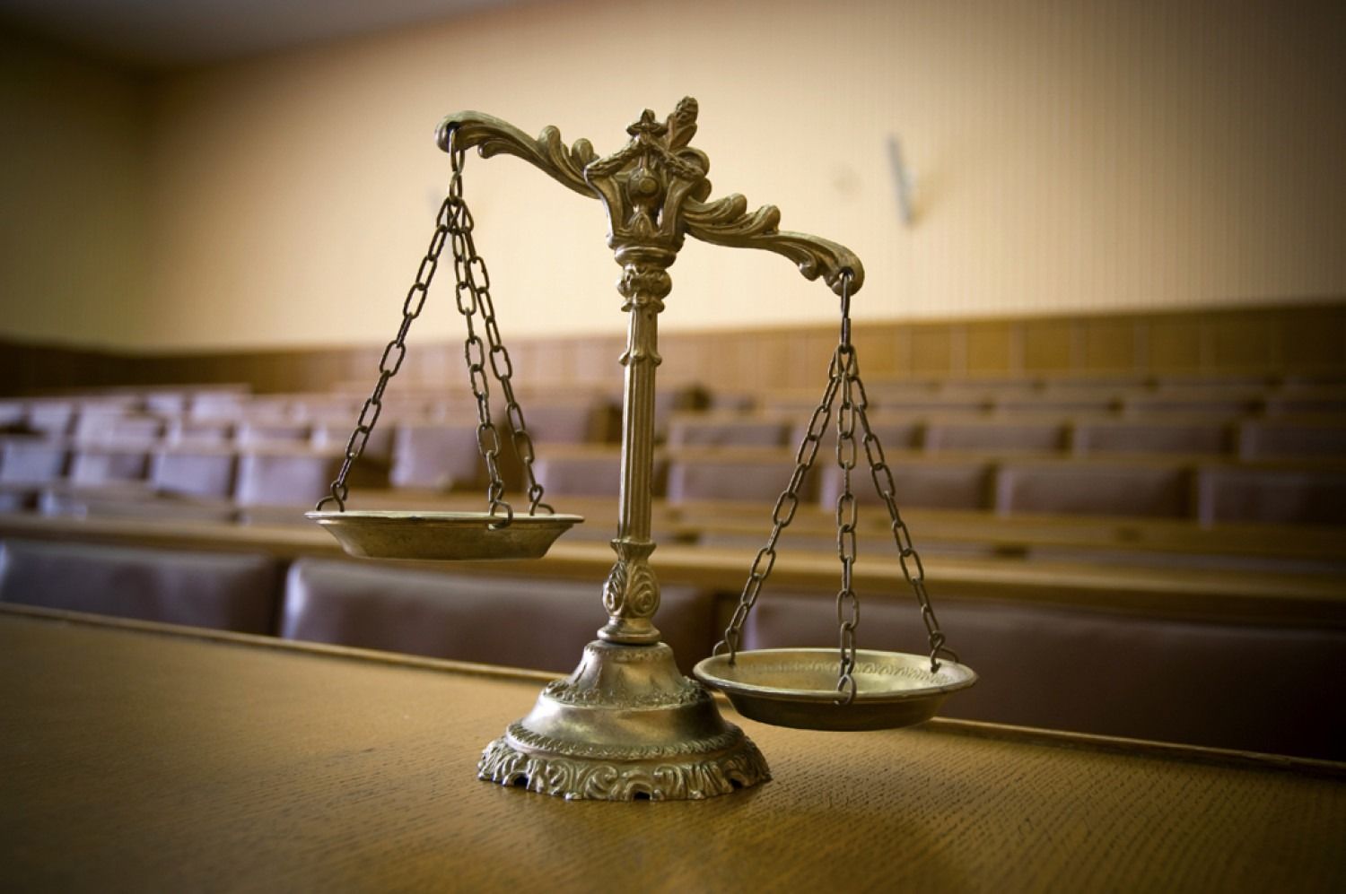 Scales of justice on a courtroom desk with rows of empty seating in the background.