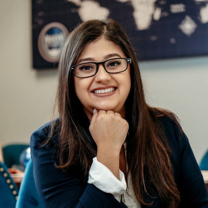 Woman with glasses, resting chin on hand, smiles at the camera; a dark blazer and a blurred world map in background.