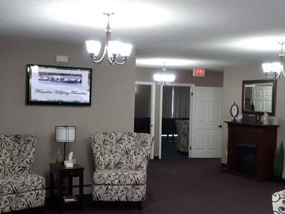 Waiting room with patterned chairs, wall TV, chandelier lights, and a hallway with a door and fireplace.
