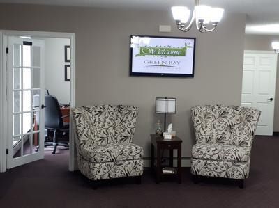Waiting area with two patterned armchairs, side table, wall sign, and open doorway to a desk.