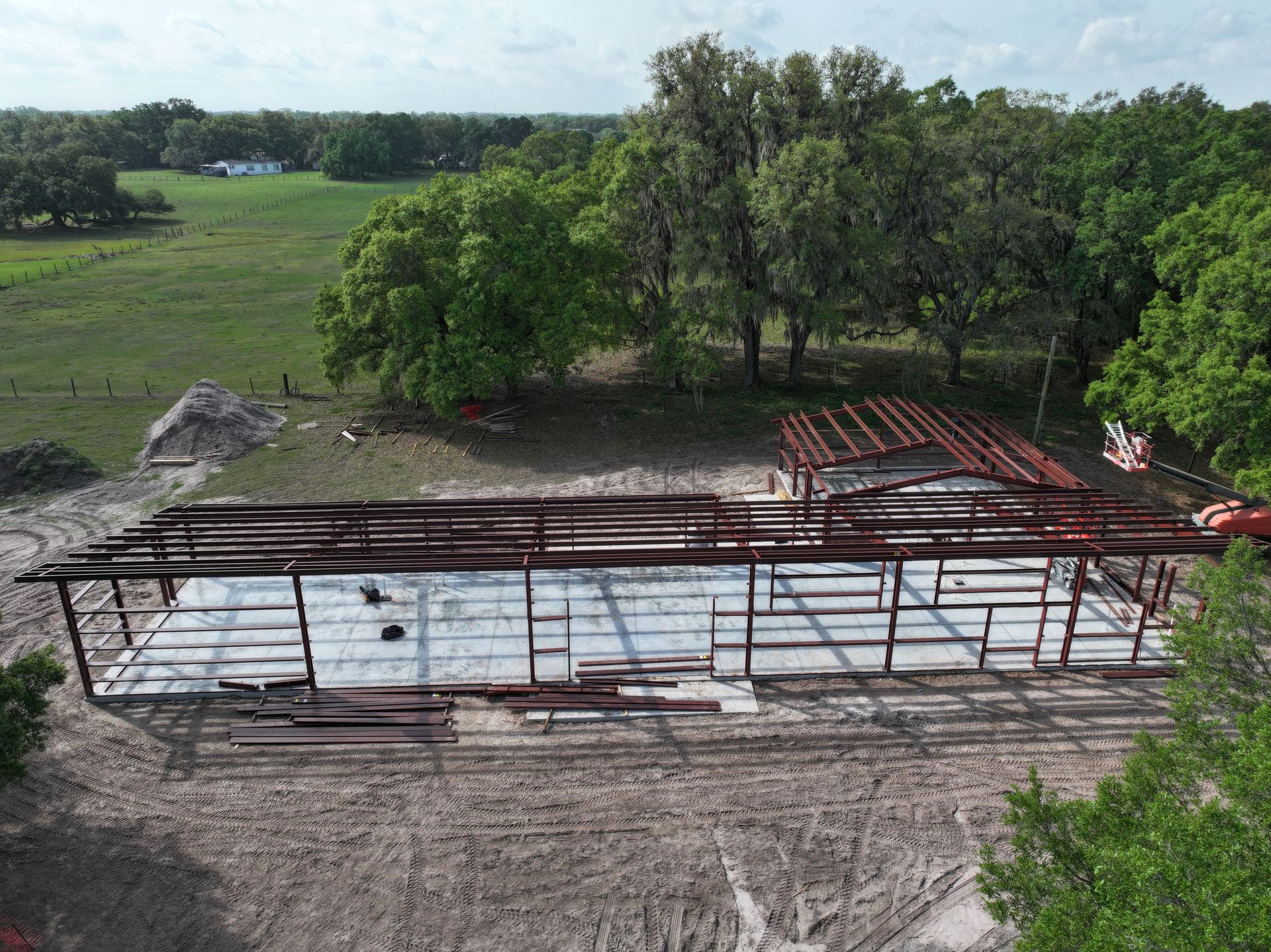 Aerial view of a metal building frame under construction in a grassy field, surrounded by trees.