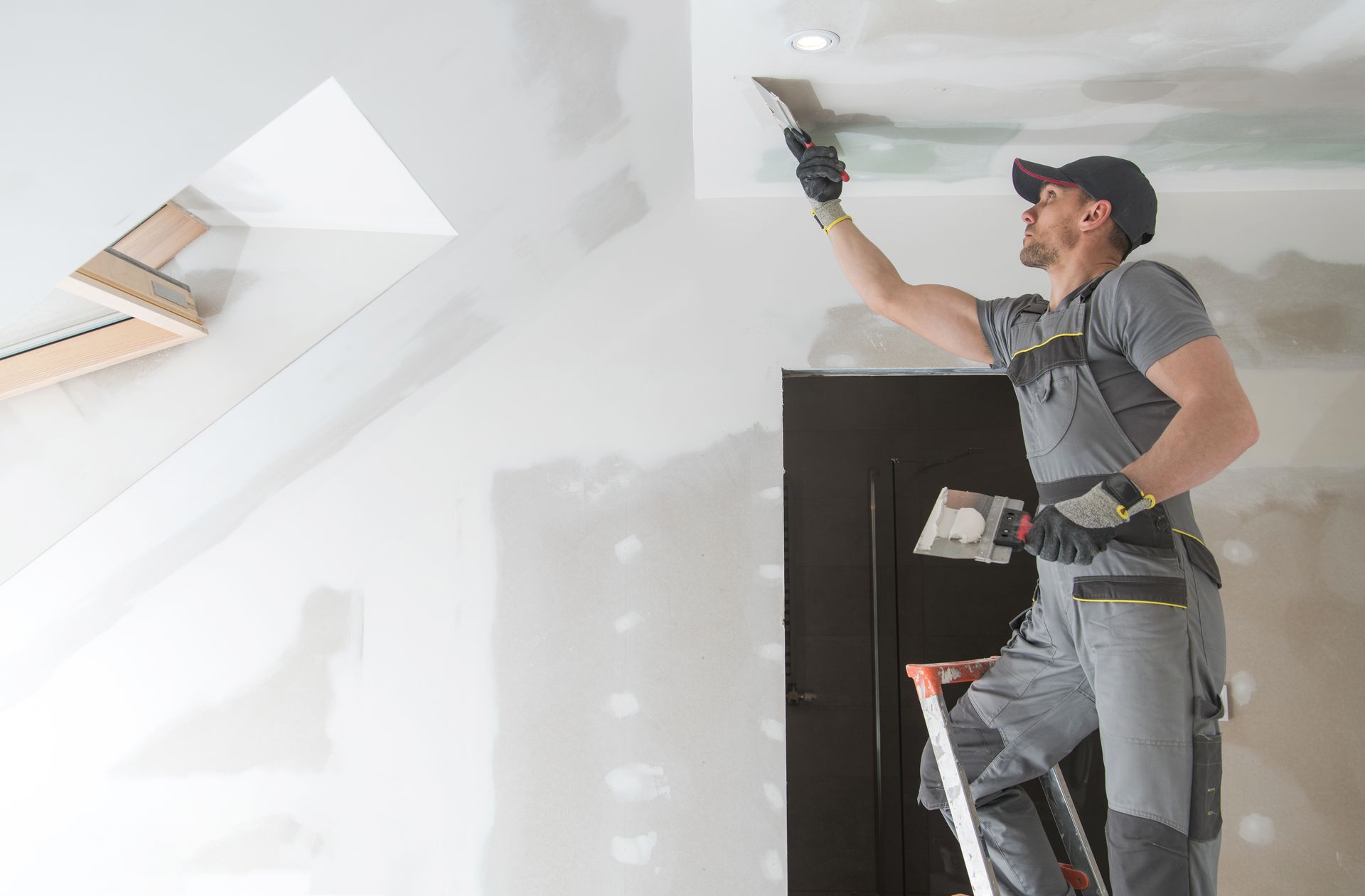 Construction worker on a ladder, applying drywall compound to a ceiling.