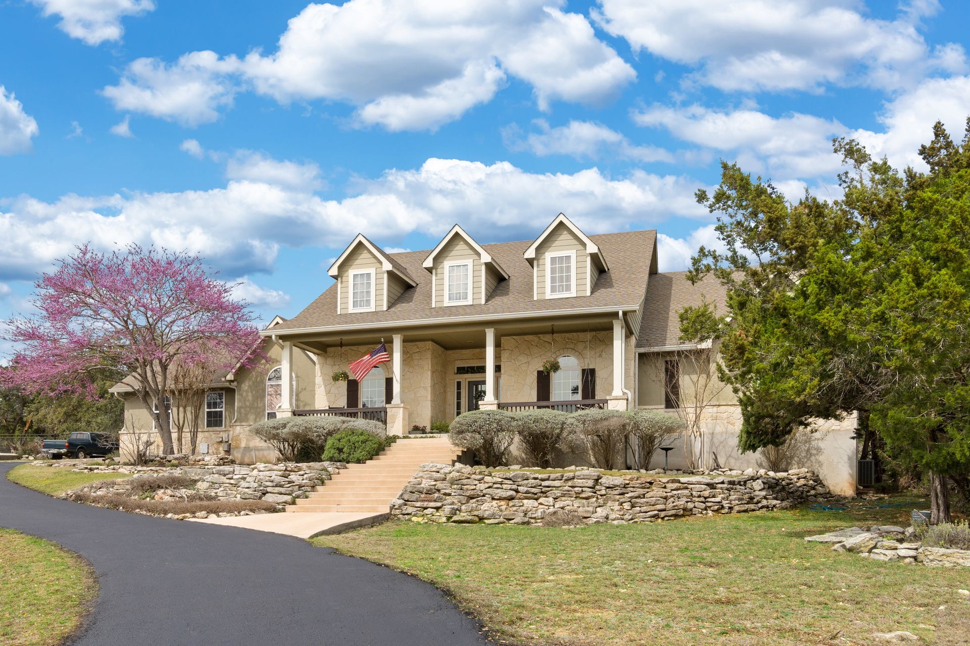 Stone house with covered porch, dormers, and curved driveway under blue sky. Stone house with covered porch, dormers, and curved driveway under blue sky.