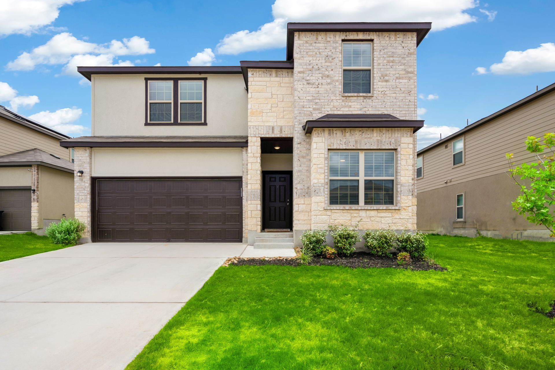 Two-story house with tan stucco and brick facade, brown garage door, and green lawn on a sunny day. Two-story house with tan stucco and brick facade, brown garage door, and green lawn on a sunny day.