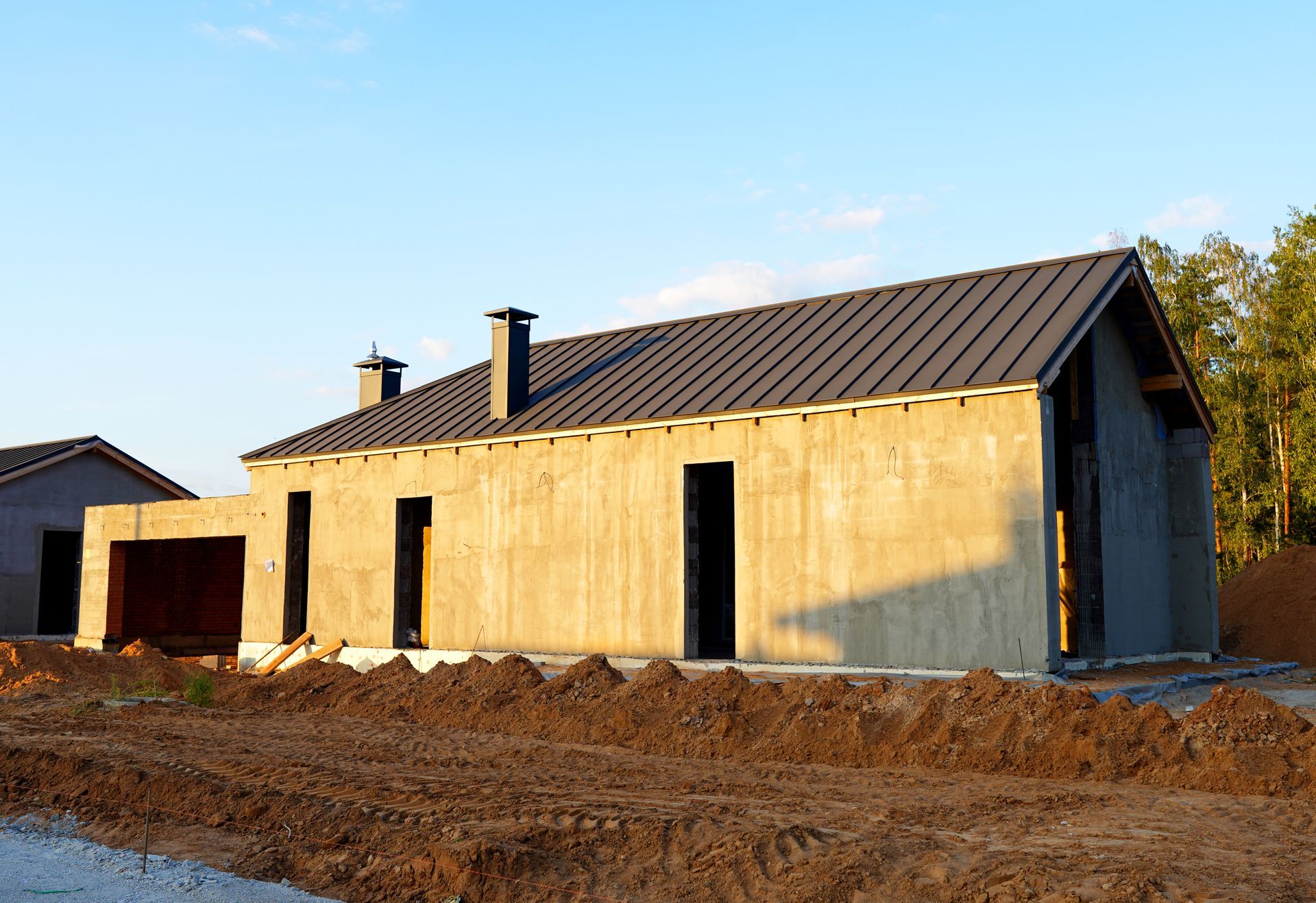 House under construction with exposed concrete walls, black roof, and dirt foreground.