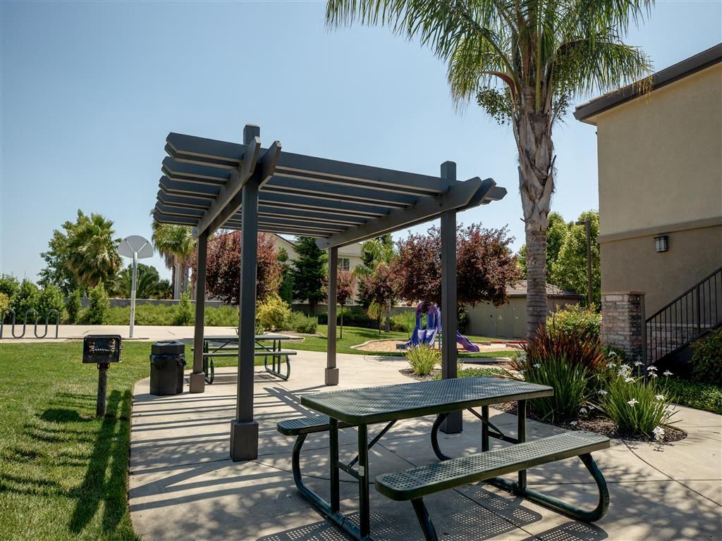 Outdoor community space with a gray pergola, picnic table, and landscaped area near a playground at Wolf Ranch Apartments.