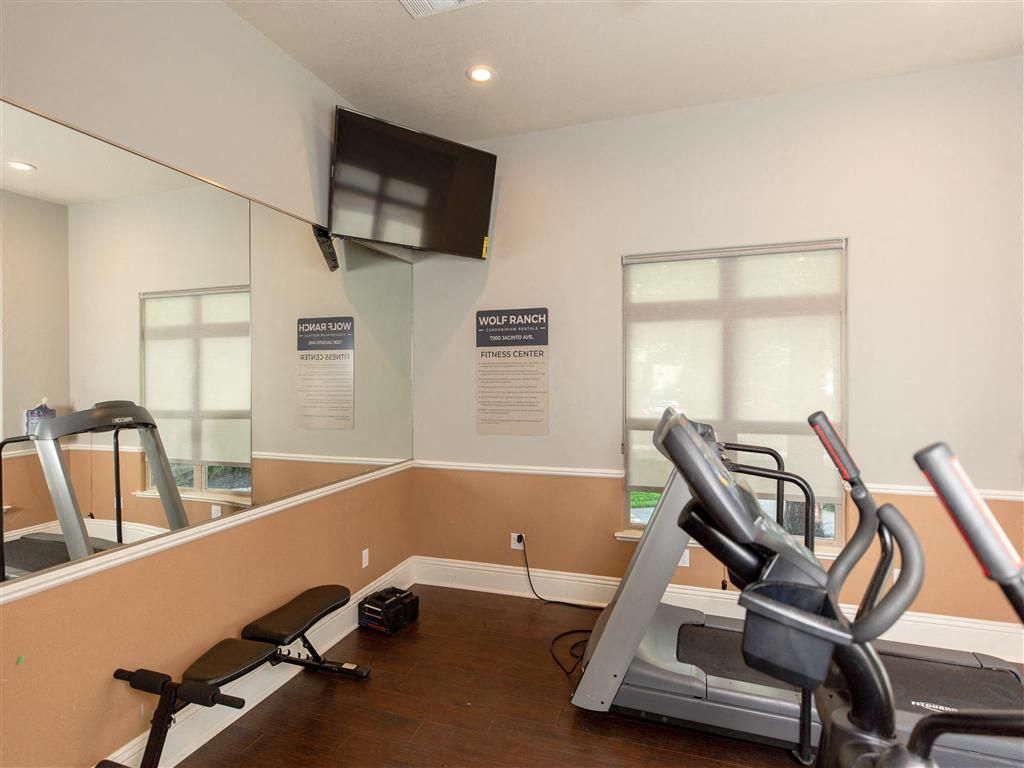 Interior of a compact community gym with treadmills, a bench, mirrors, and a wall TV at Wolf Ranch Apartments at Wolf Ranch Apartments.