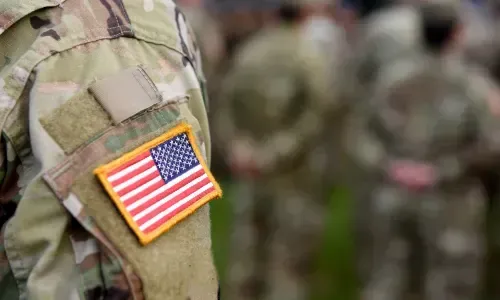 US flag patch on a military uniform sleeve, other soldiers in the blurry background.