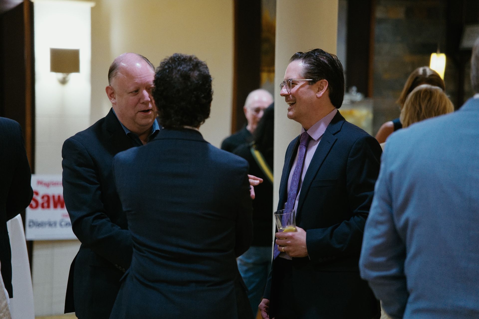 Three men talking in a hotel lobby, one in a dark suit holding a drink.