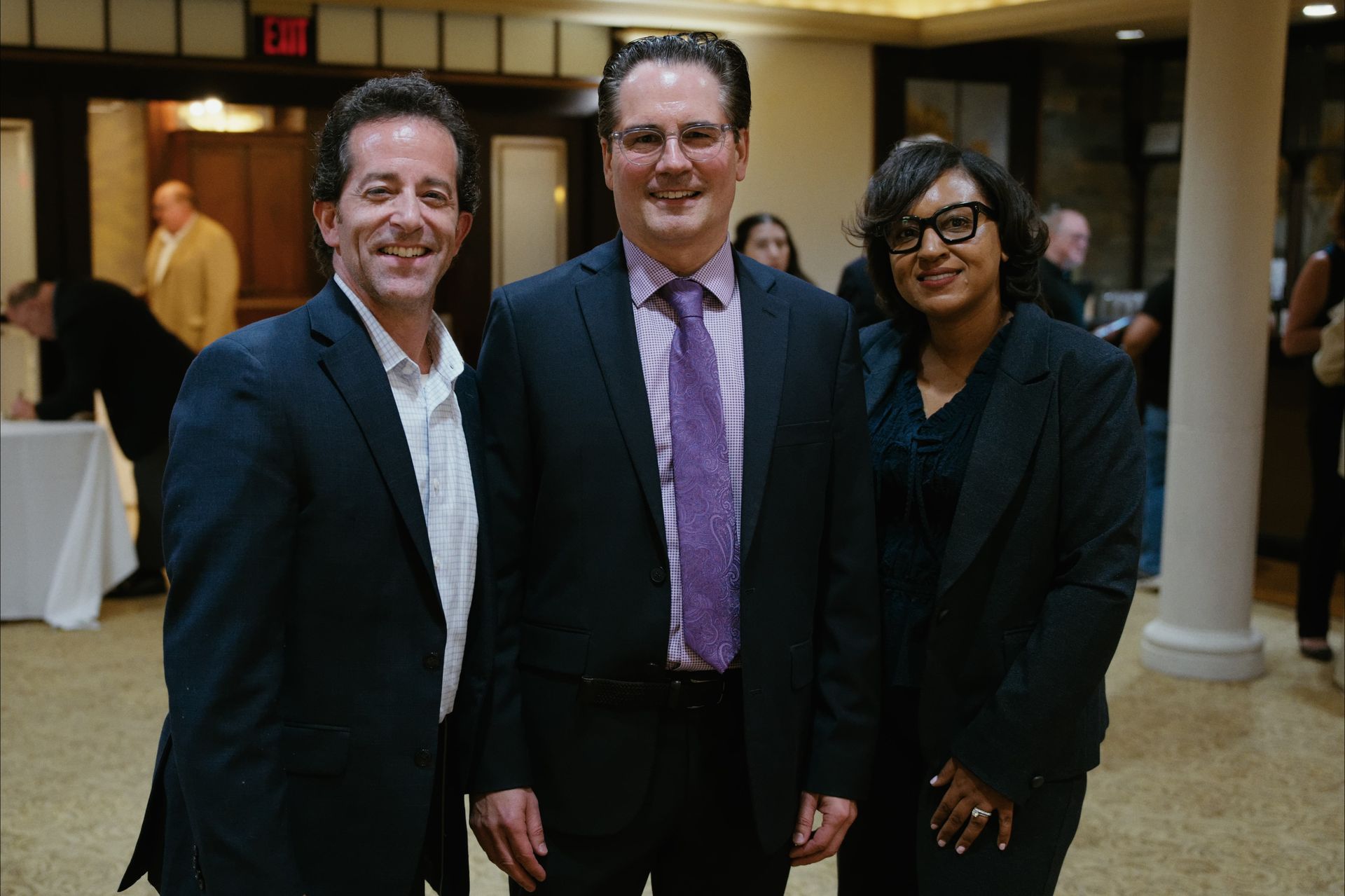 Three people in suits posing indoors at an event, smiling in a lobby-like space.