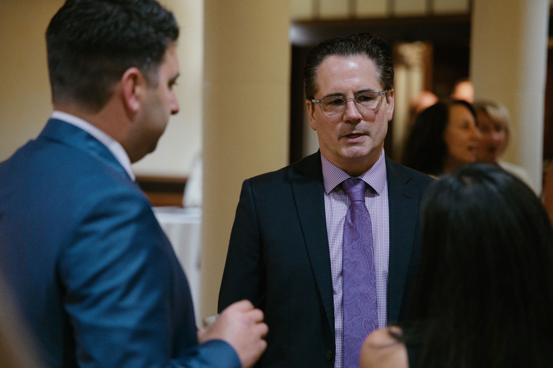 Two men in suits converse in a hallway, one facing the camera while others blur in the background.