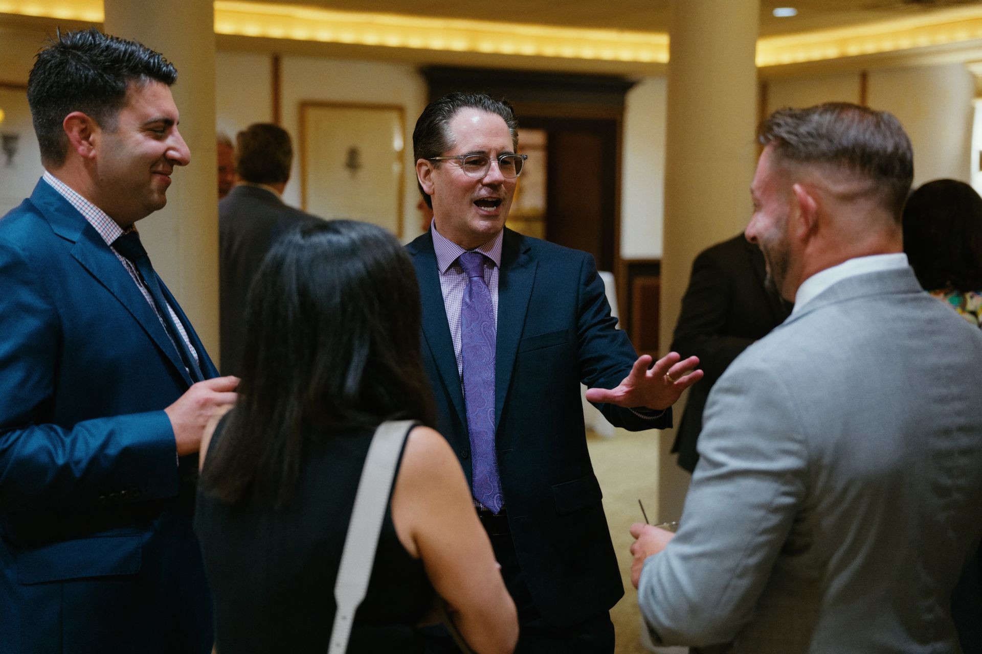Four people in business attire talking in a warmly lit indoor lobby or conference setting.